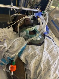 Keith in a hospital bed wearing a neck brace, connected to medical lines