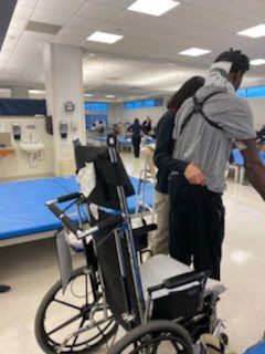 Keith standing with help in a rehab gym, a wheelchair beside him